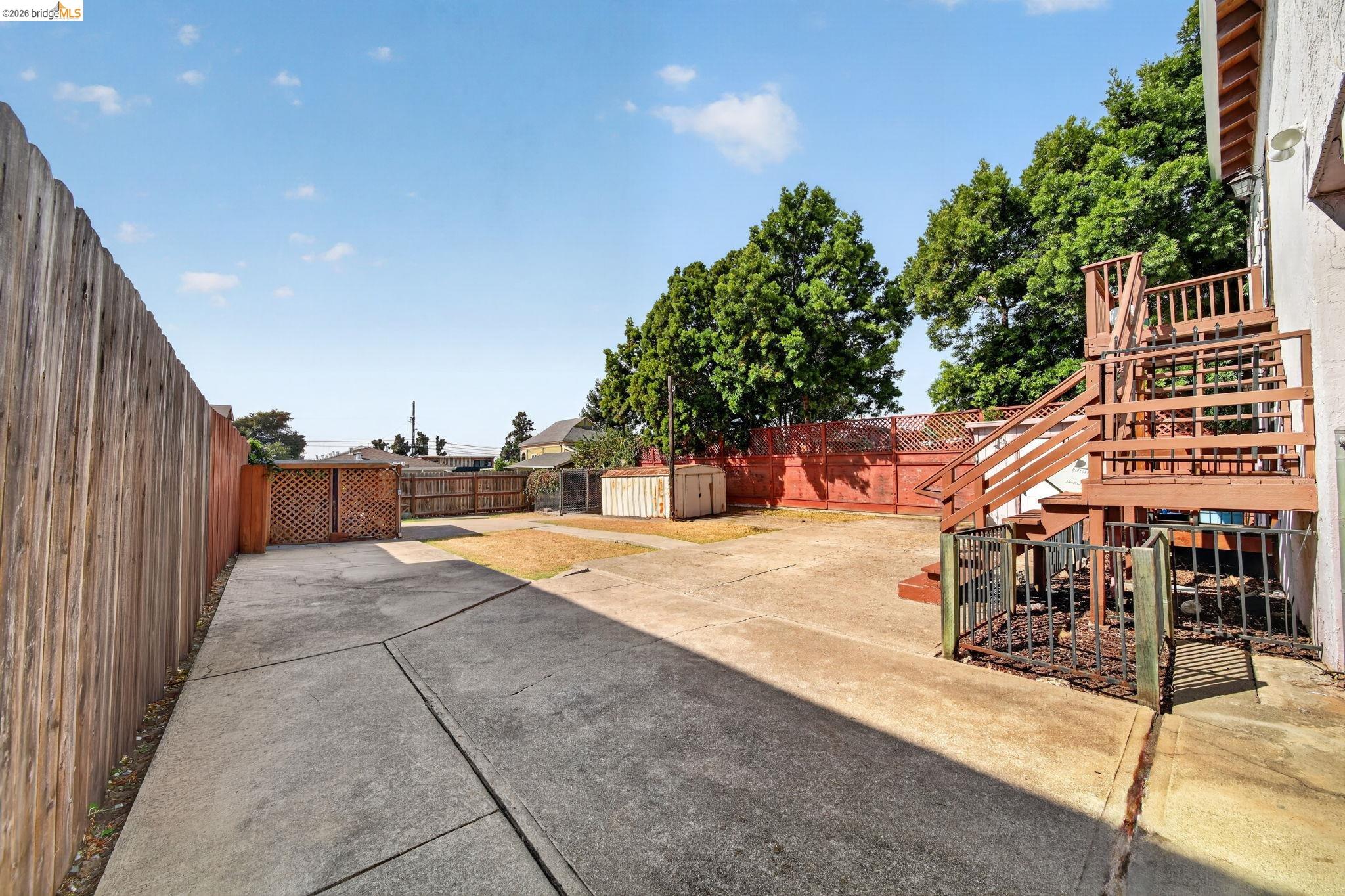Fenced backyard with a broad concrete patio, wooden deck and stairs, mature trees, and a storage shed.