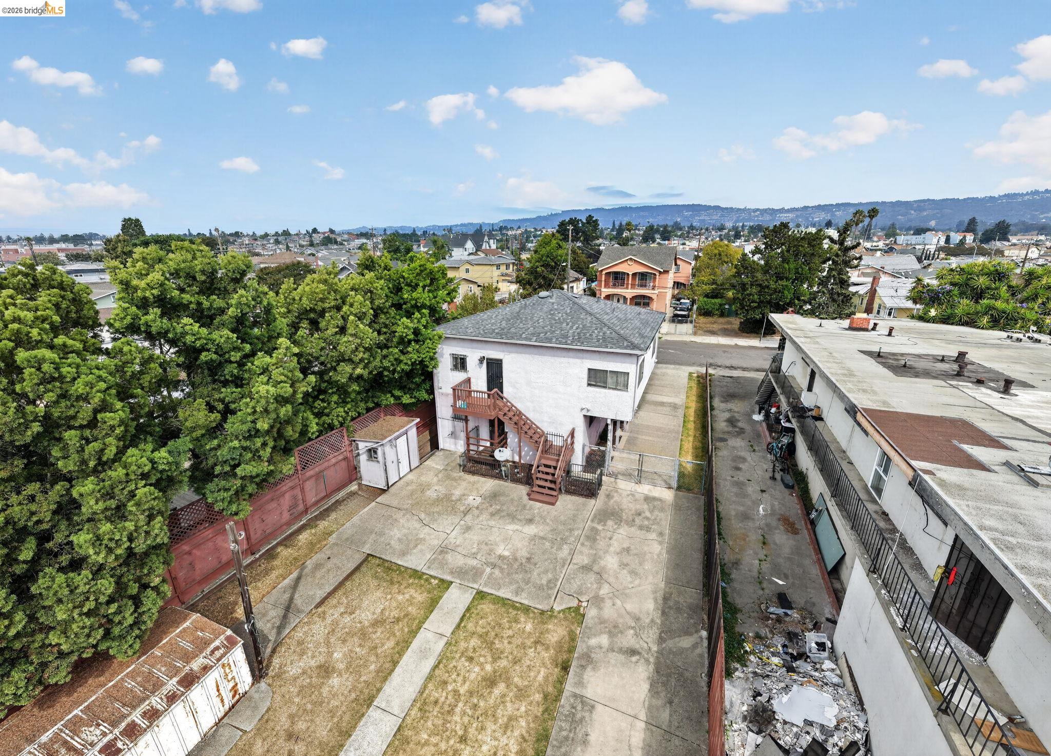 Aerial view of a white residence’s rear, concrete yard, mature trees, and surrounding Oakland neighborhood.