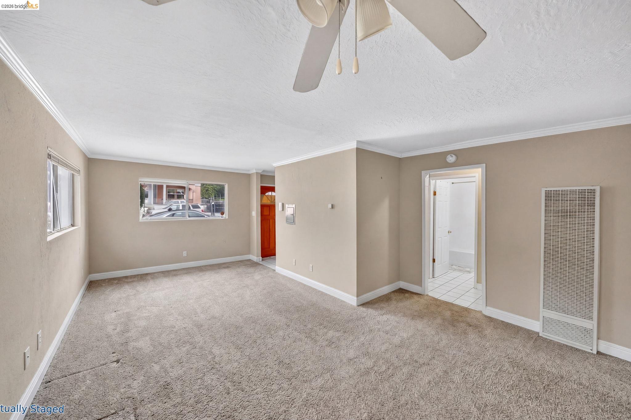 Empty living room with beige walls, carpet, ceiling fan, and windows.