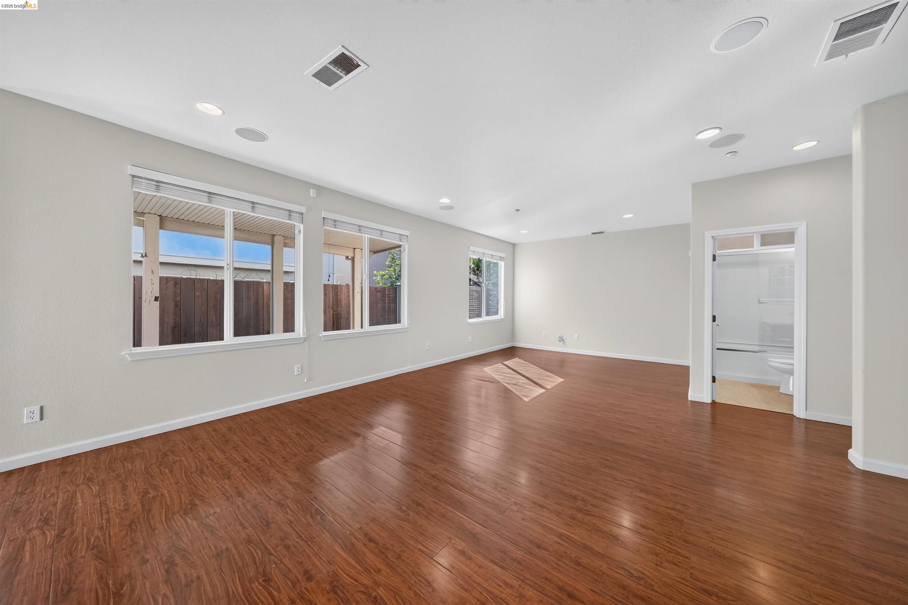 Empty living room with warm wood-look flooring, broad windows, and an adjoining bathroom.