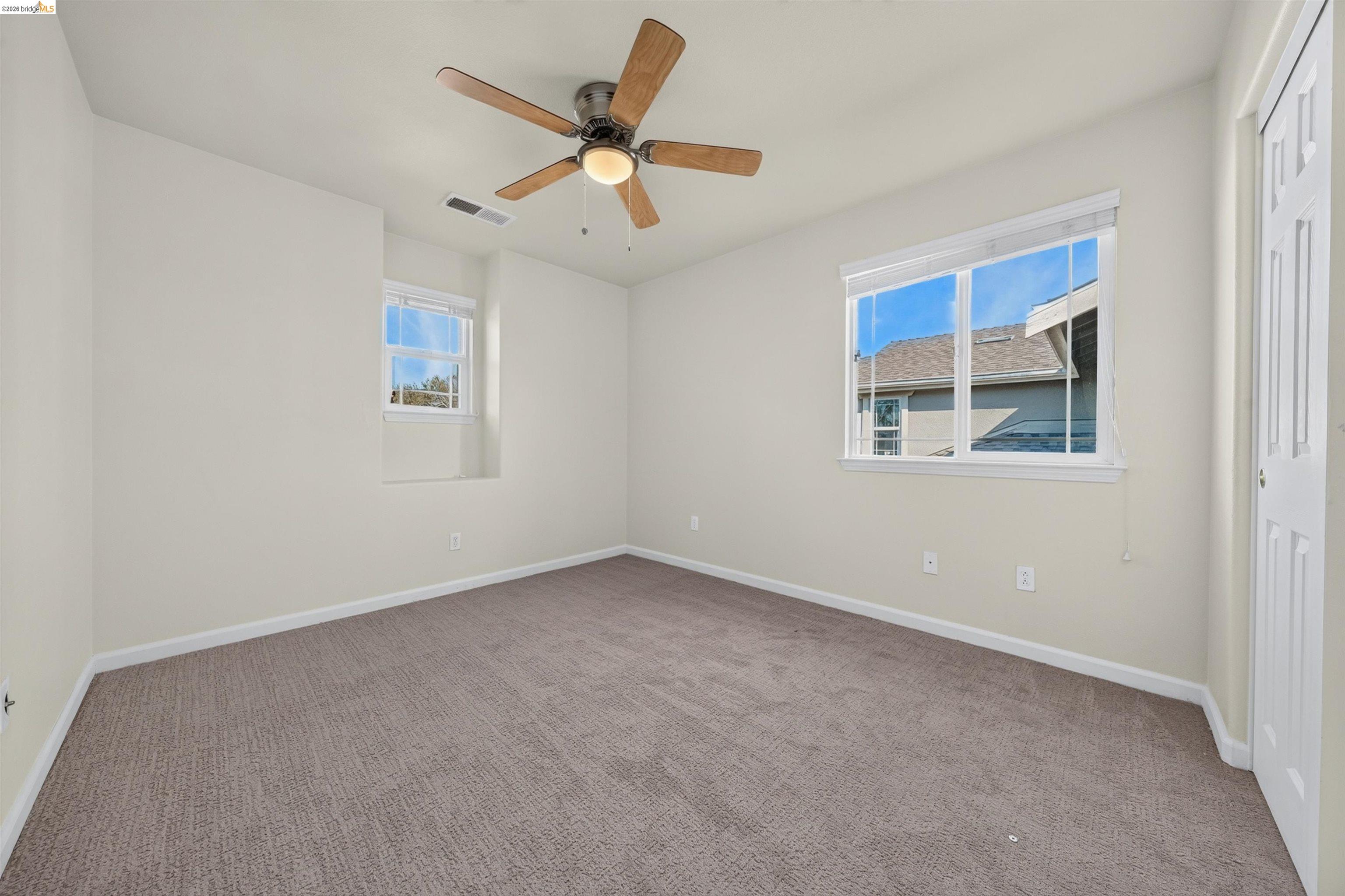 Empty carpeted bedroom with a ceiling fan and two windows.