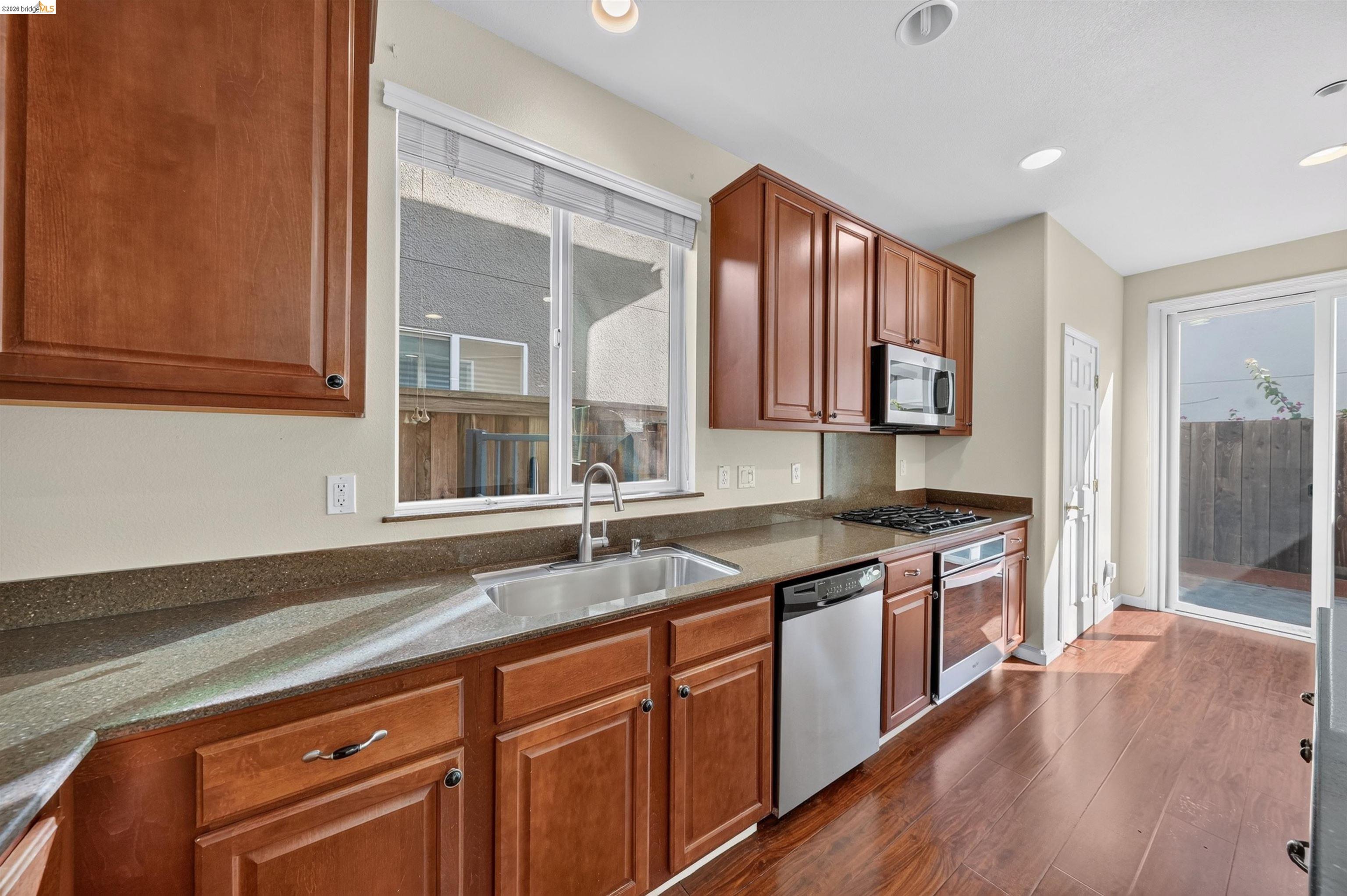Kitchen with reddish wood cabinets, granite counters, stainless appliances, and a window overlooking a fenced yard.