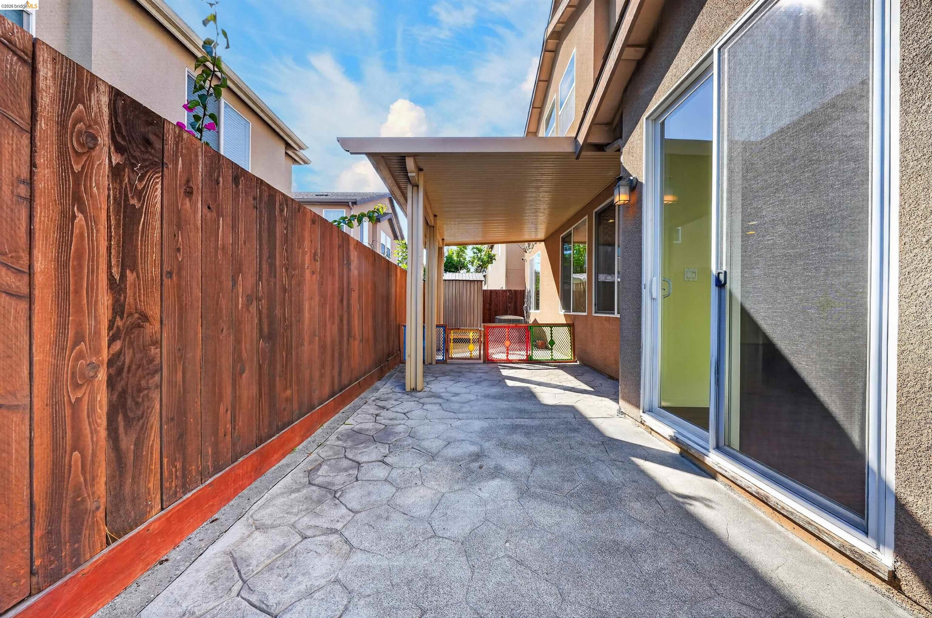 Covered side patio with stamped concrete paving, a tall wooden fence, and sliding glass doors.