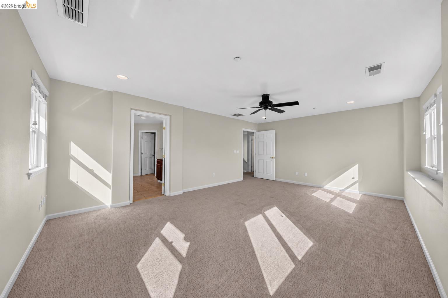 Empty carpeted bedroom with a ceiling fan, windows, and sunlight across the floor.