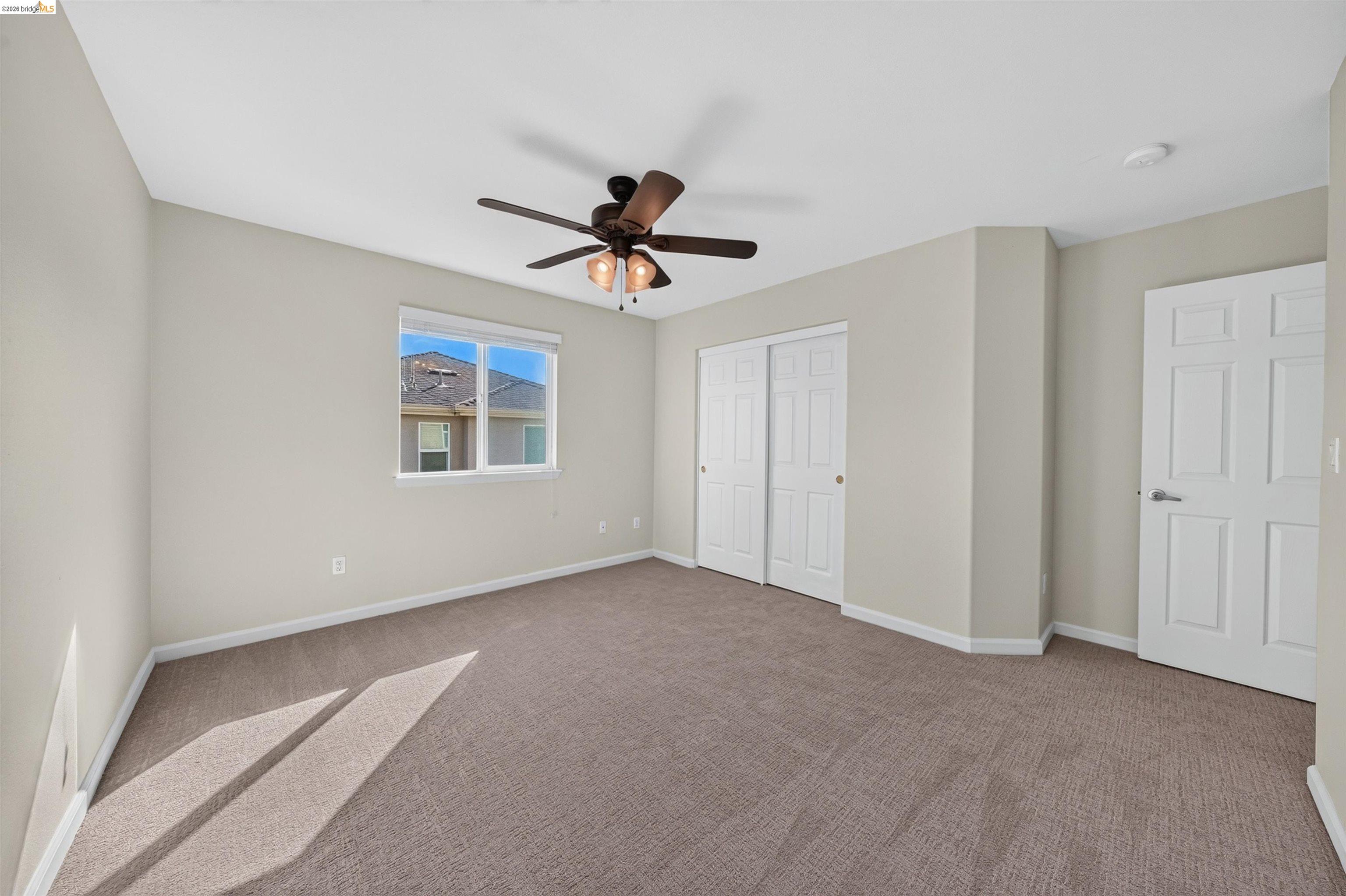 Empty carpeted bedroom with a ceiling fan, window, and double closet doors.