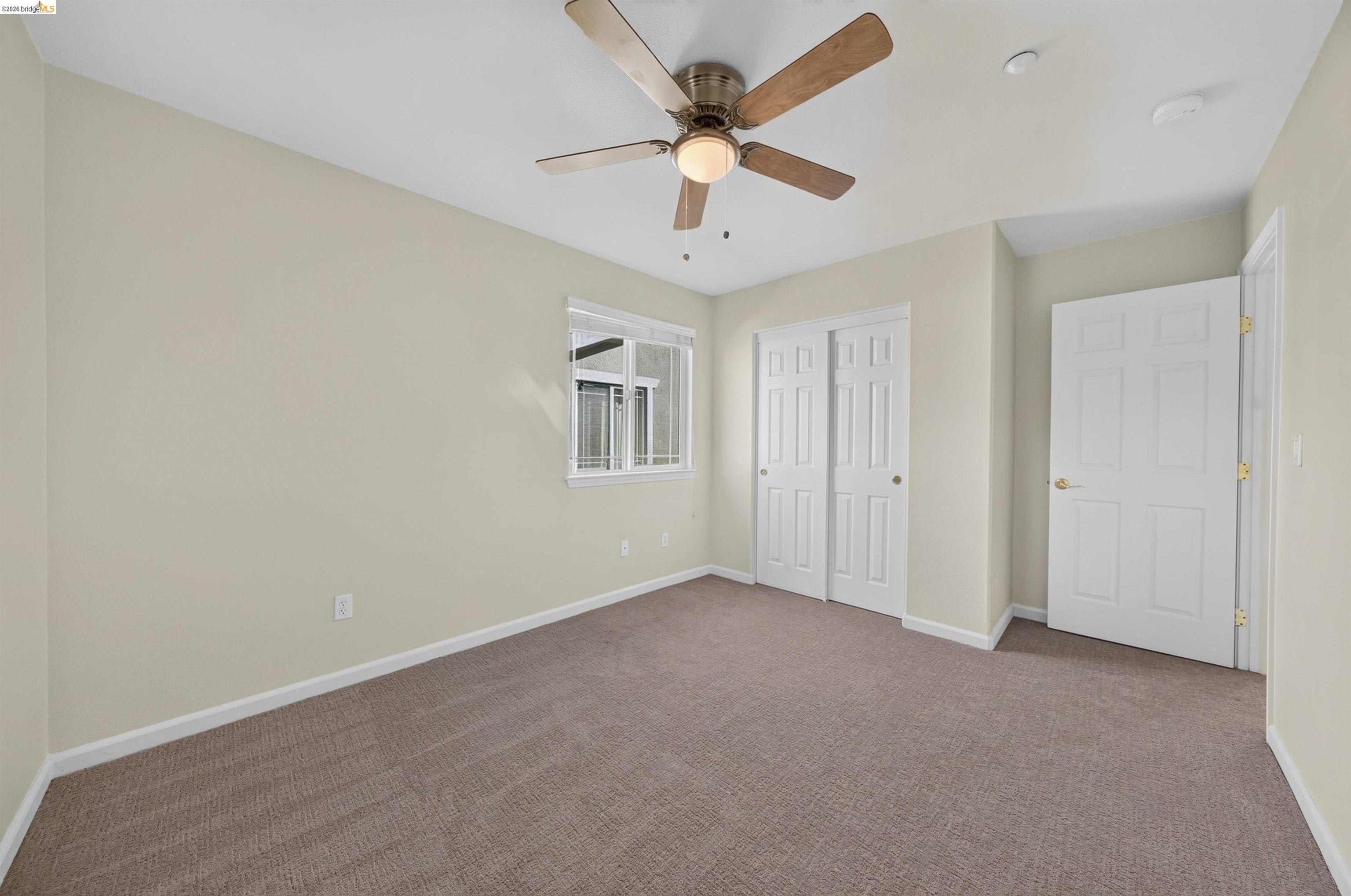 Empty carpeted bedroom with a ceiling fan, window, and double closet doors.