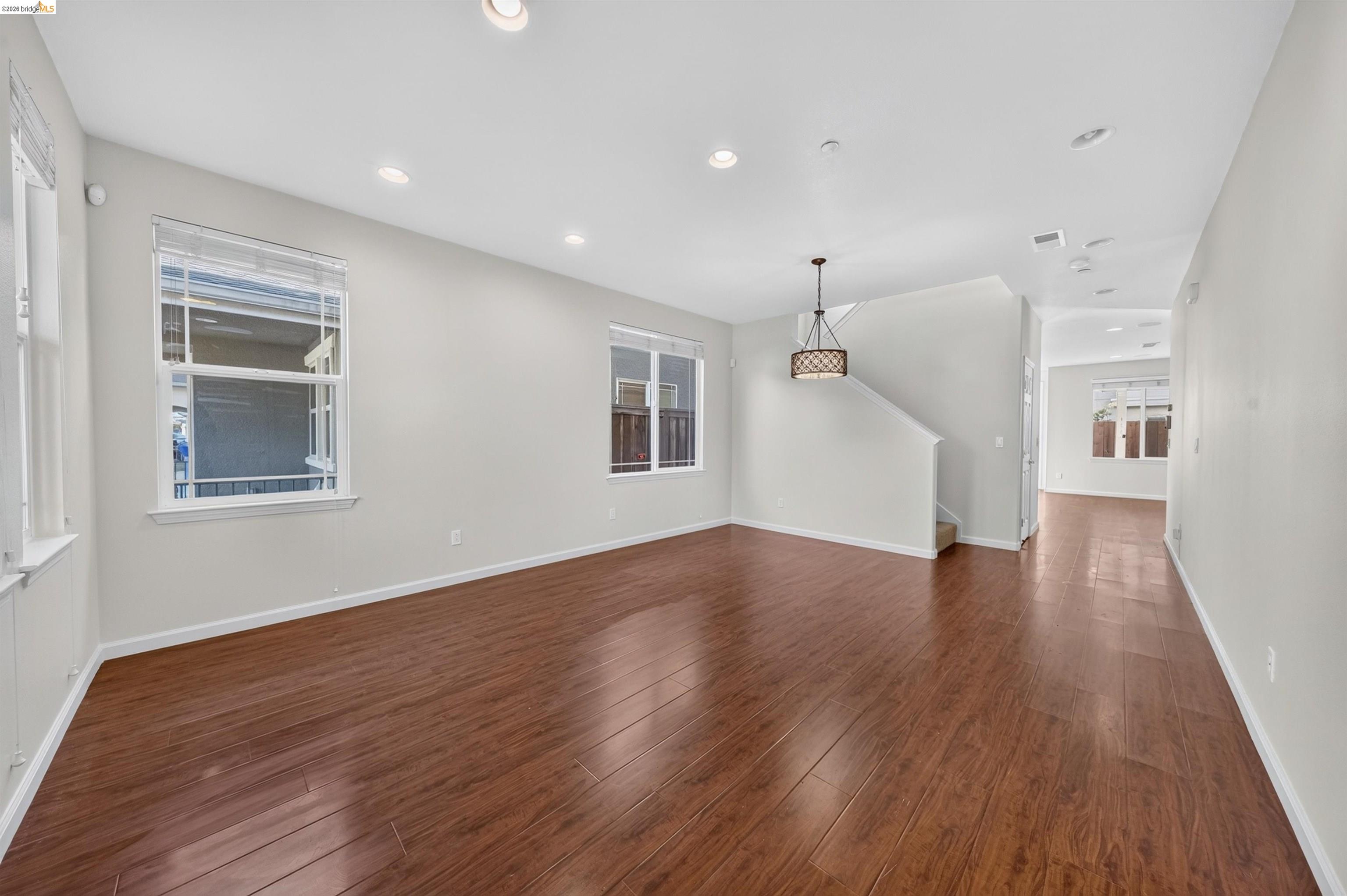 Empty living room with reddish-brown plank flooring, large windows, and a staircase.