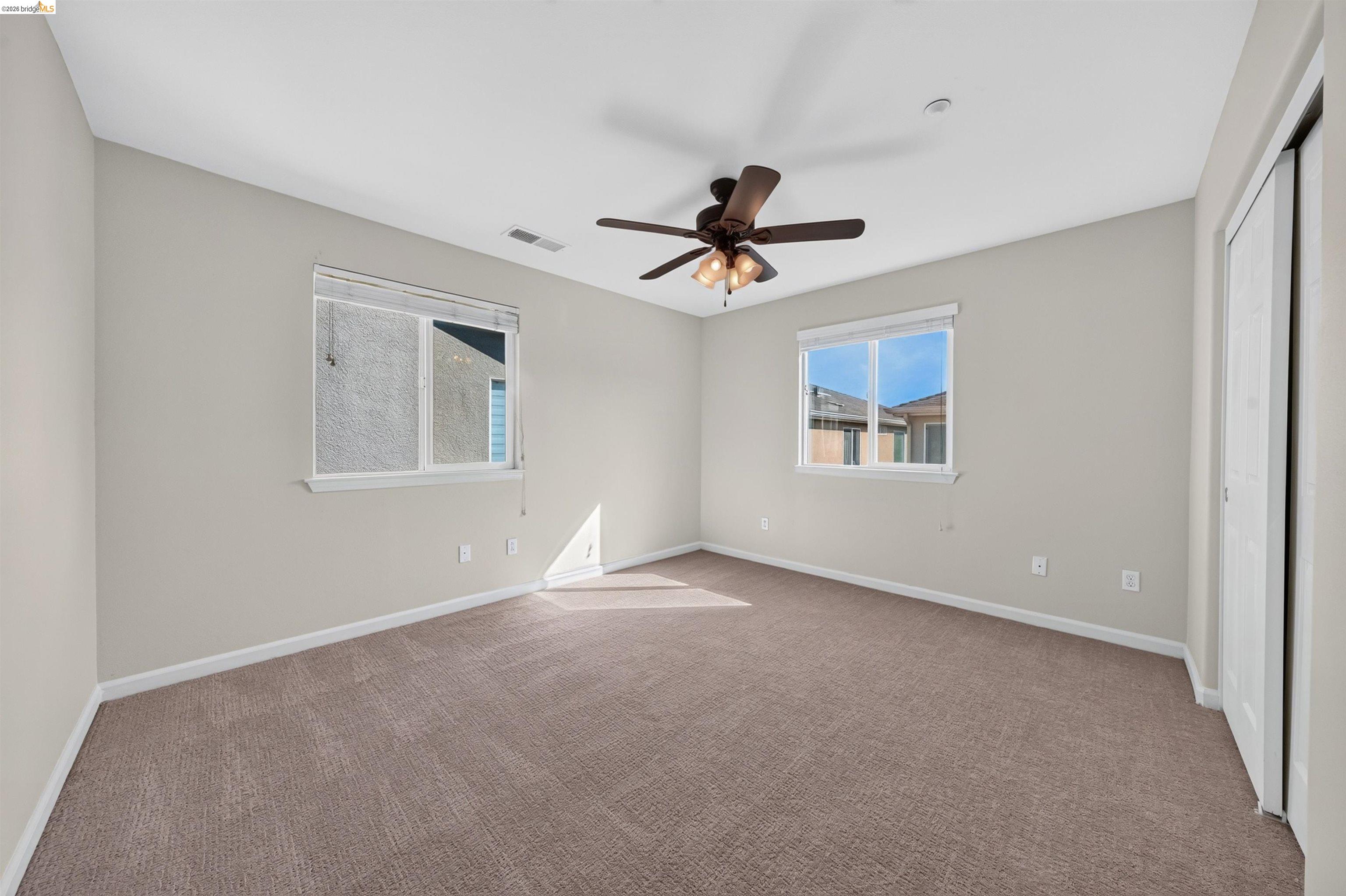 Empty bedroom with beige carpet, two windows, and a ceiling fan.