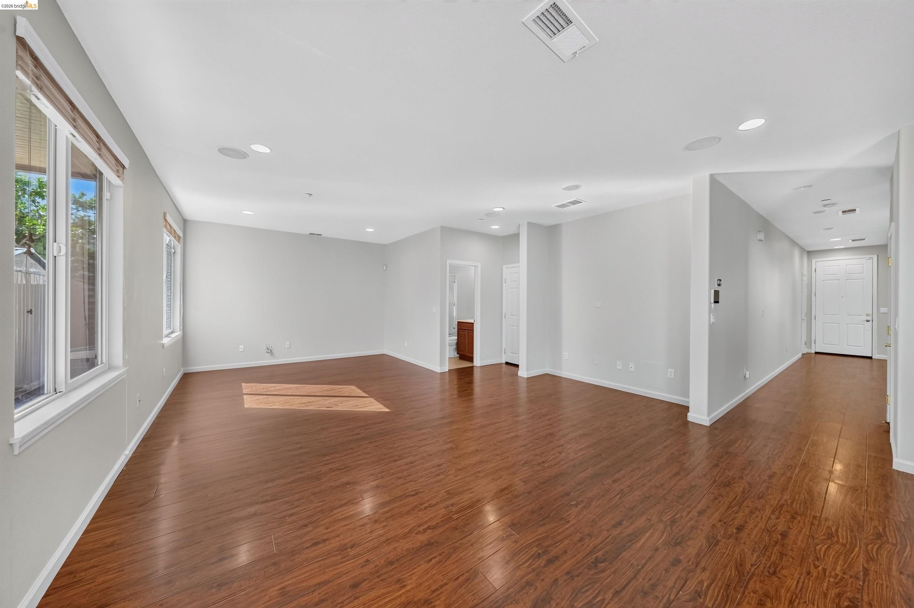 Empty living area with warm wood flooring, large windows, and an entry hallway.
