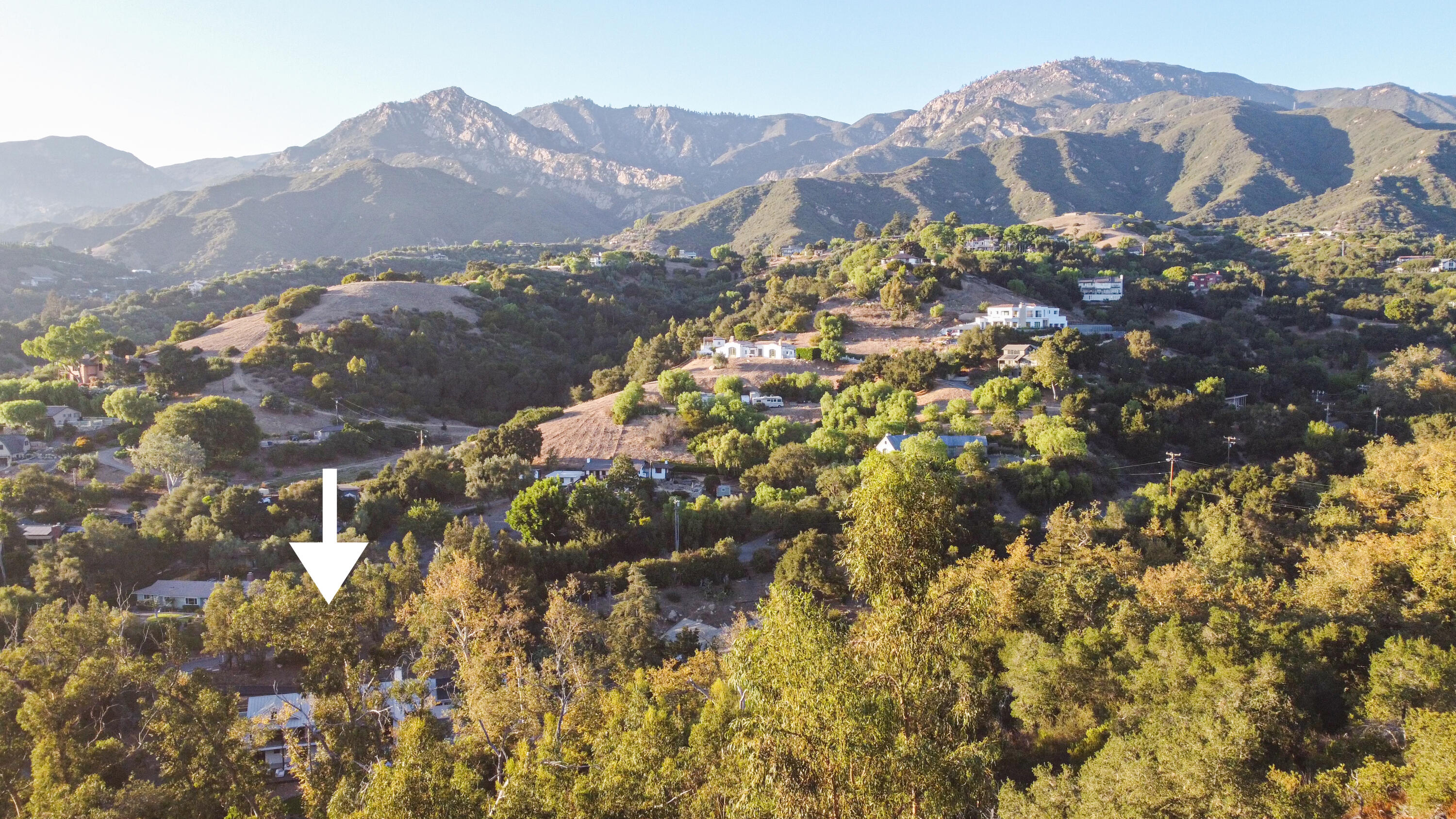 Aerial hillside property marked by a white arrow, with wooded surroundings and mountain views.