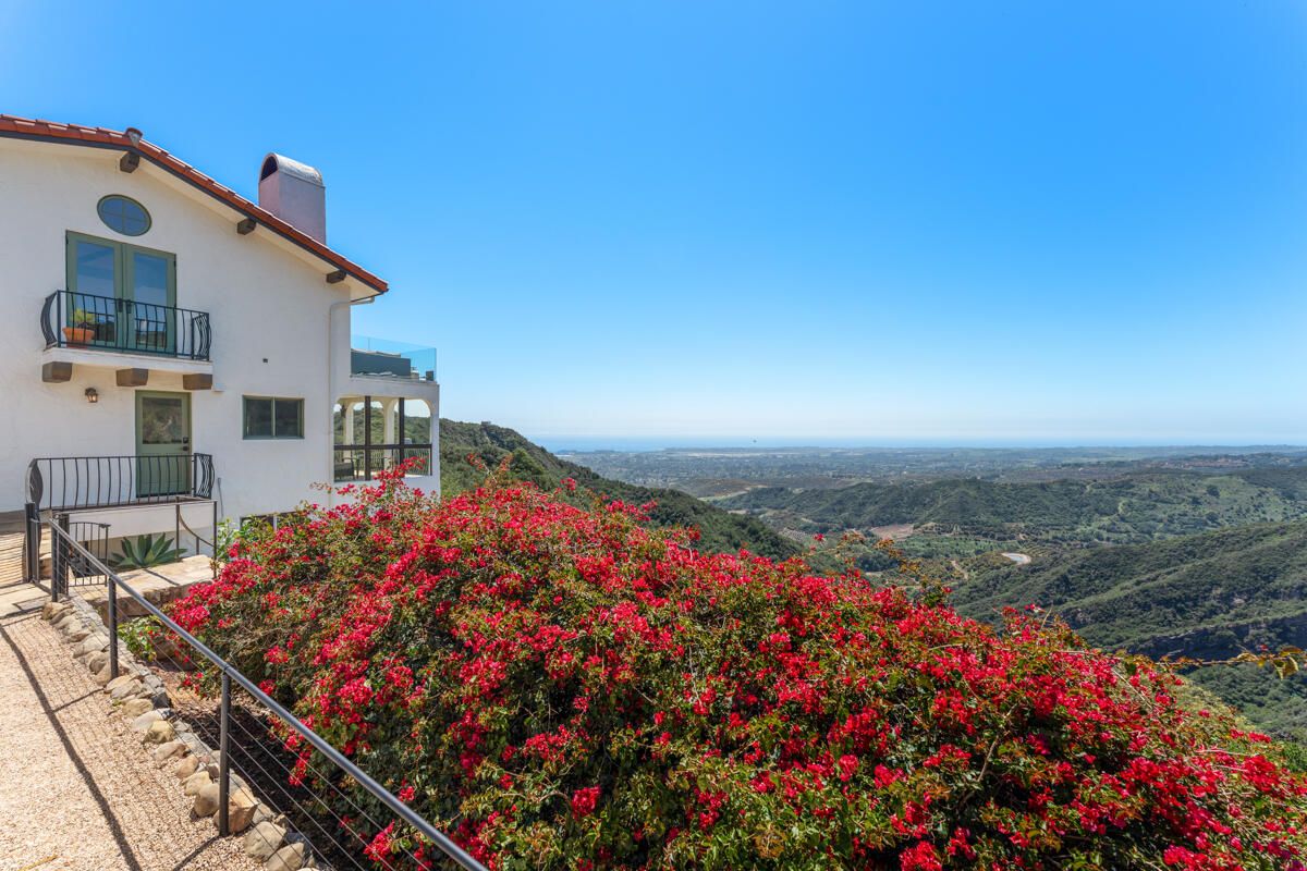 White stucco home with balconies overlooks flowering shrubs, green hills, and the distant ocean.