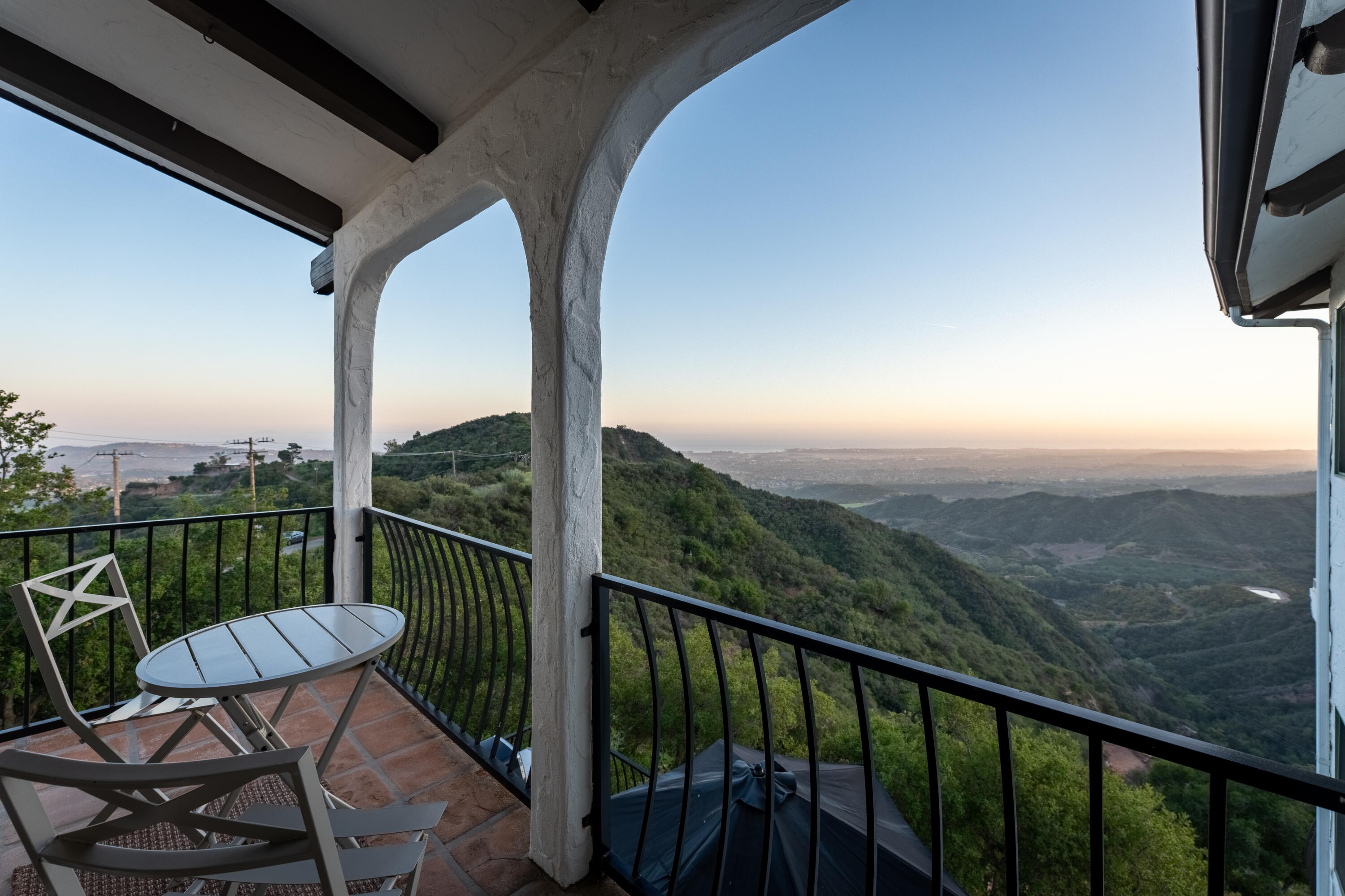 Covered balcony with stucco arches and patio seating overlooking green hills and the distant coast at golden hour.
