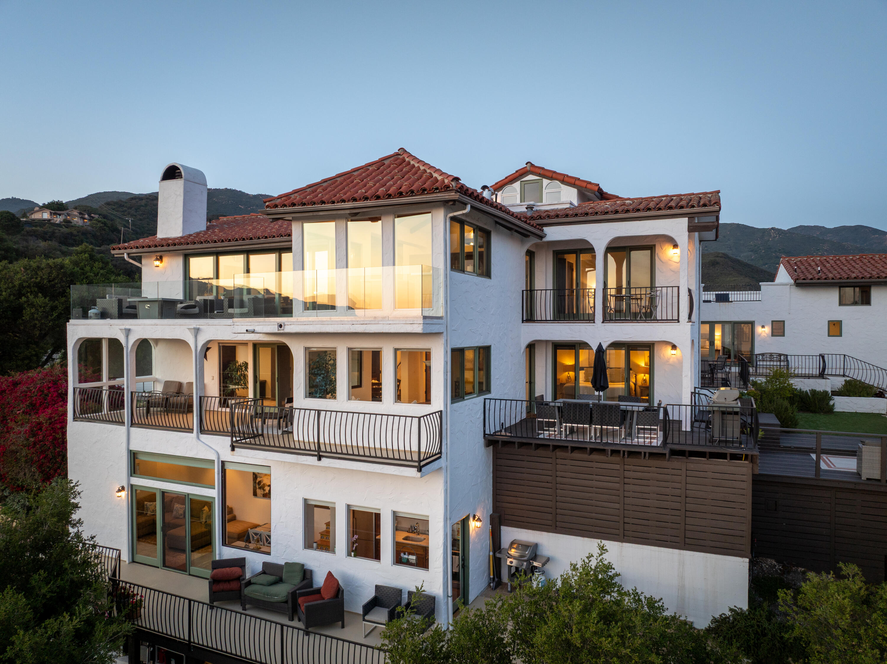 White stucco home with terracotta tile roofs, multiple balconies, and warmly lit rooms at dusk.