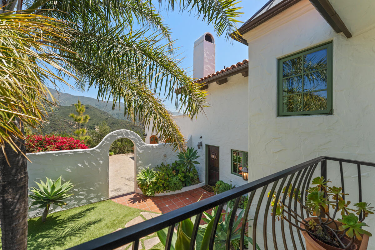 White stucco home beside a tiled courtyard and arched garden wall, with palms and mountains beyond.