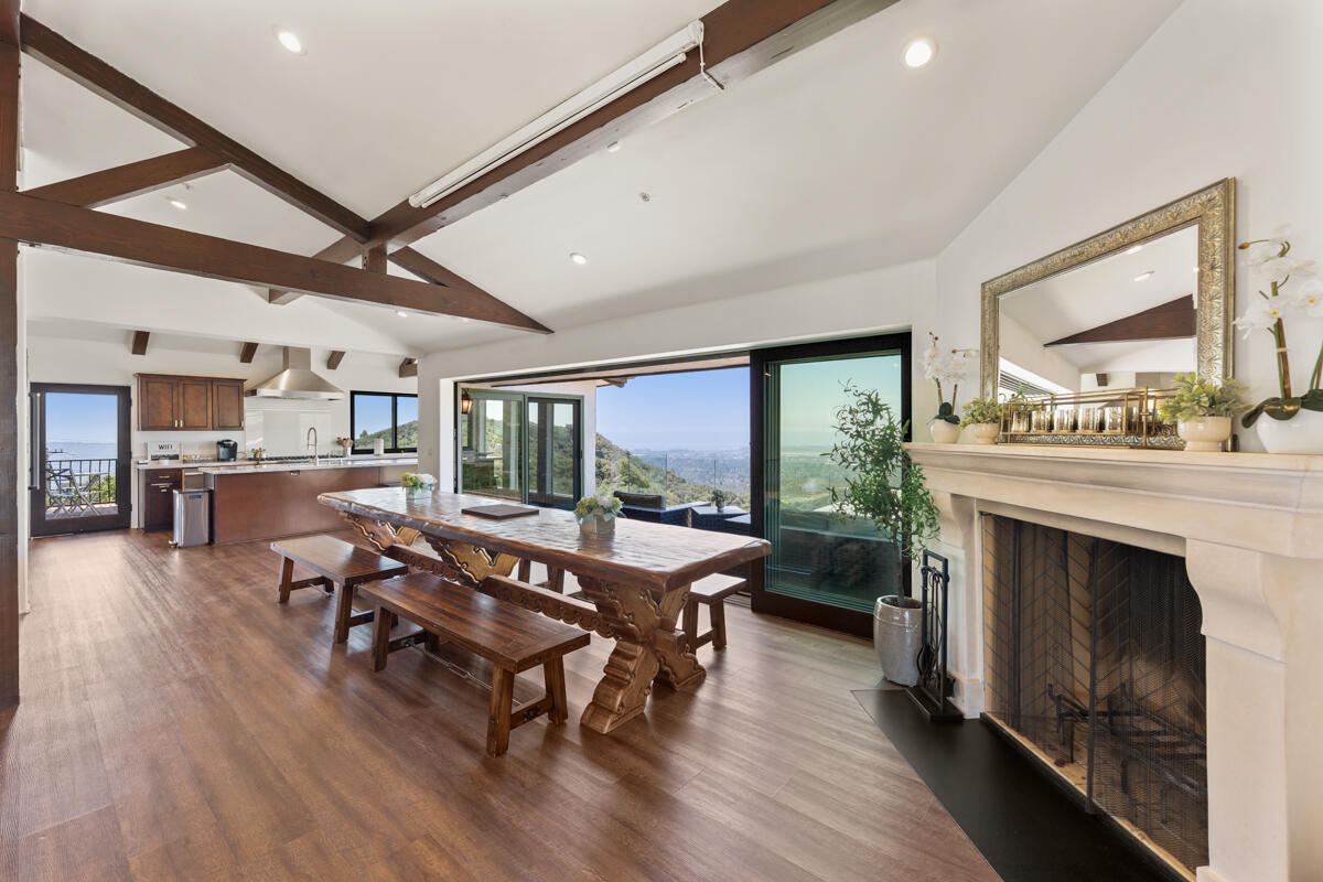 Open dining area with exposed wood beams, a carved table, stone fireplace, and hillside water view.
