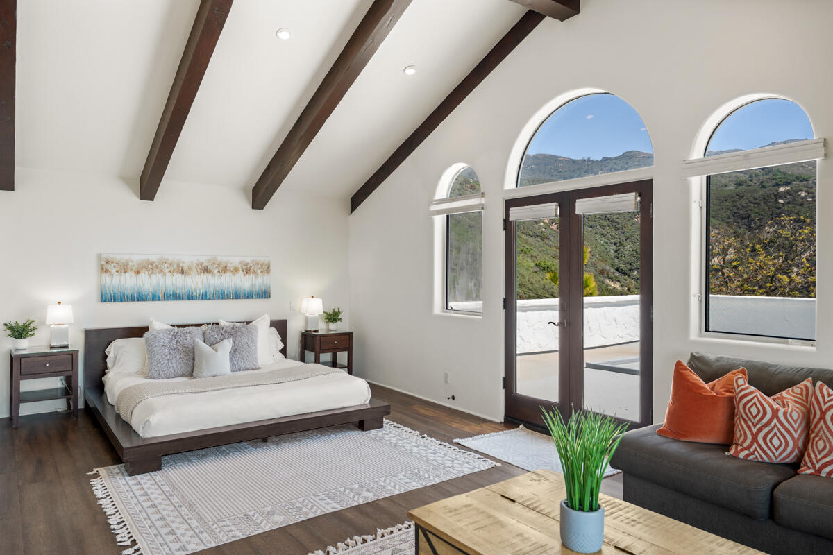 Staged bedroom with exposed ceiling beams, arched windows, and views of green hills.