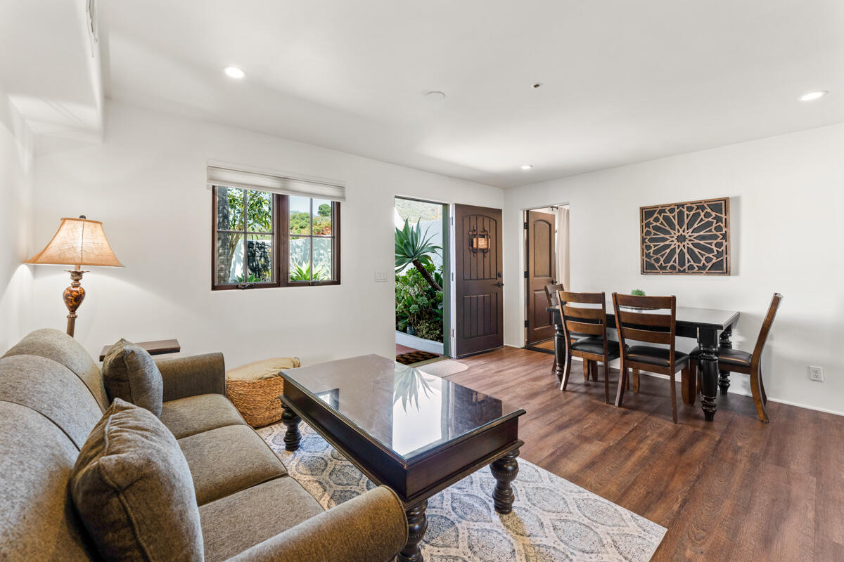 Living and dining area with a gray sectional, dark wood dining set, and open door beside garden greenery.