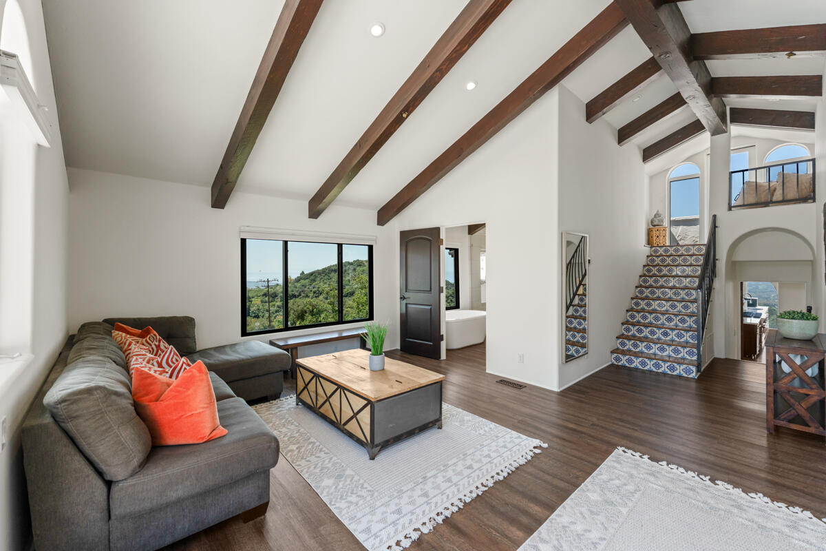 Living room with exposed ceiling beams, a gray sectional, and a blue-and-white tiled staircase.