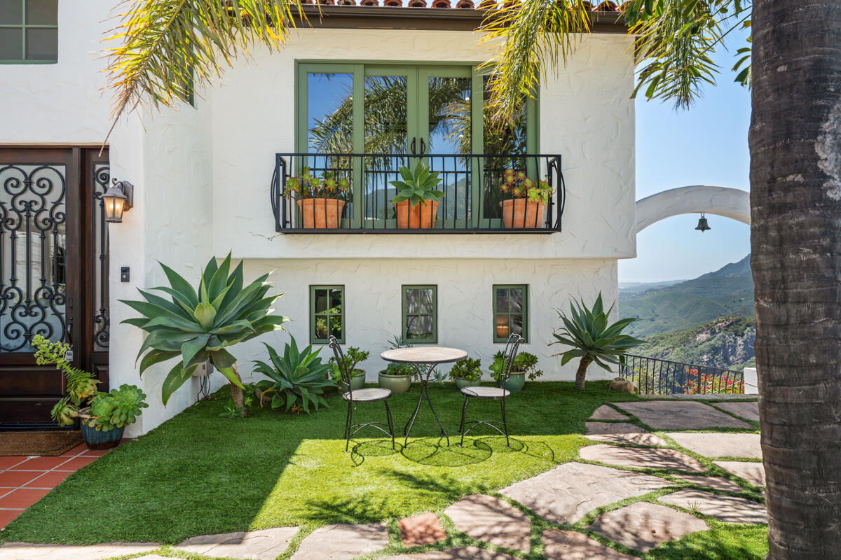 White stucco home with a green-framed balcony overlooking a landscaped lawn and distant hills.