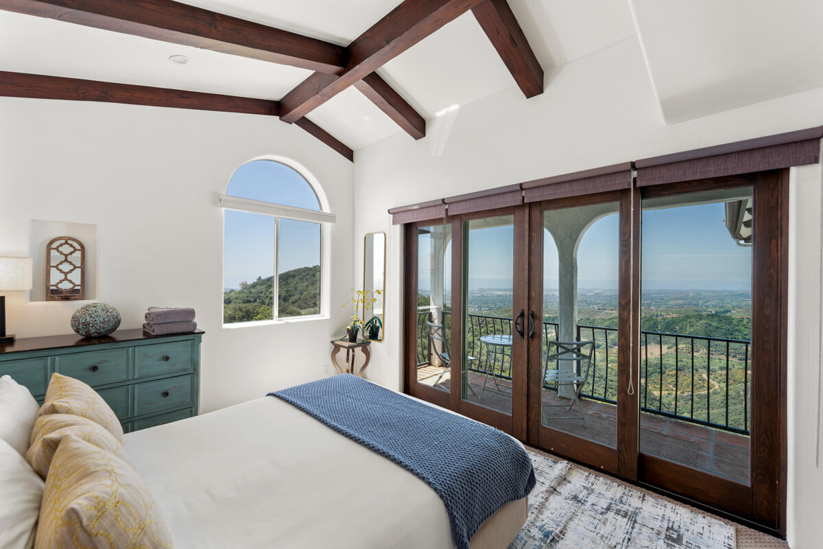 Bedroom with exposed wood beams and sliding doors to a balcony overlooking green hills and the coast.