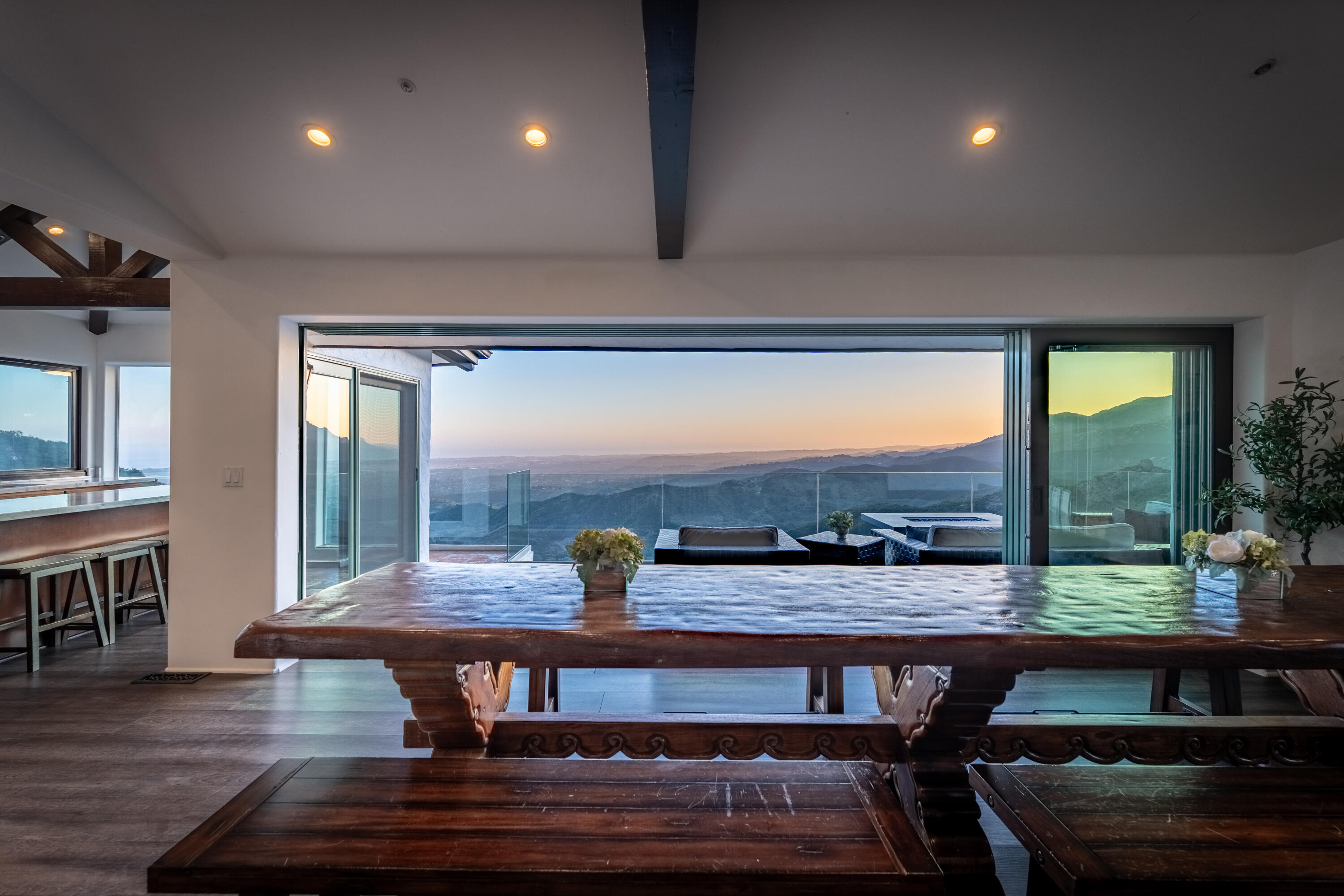 Dining room with a carved wooden table opening to a terrace and mountain views at sunset.