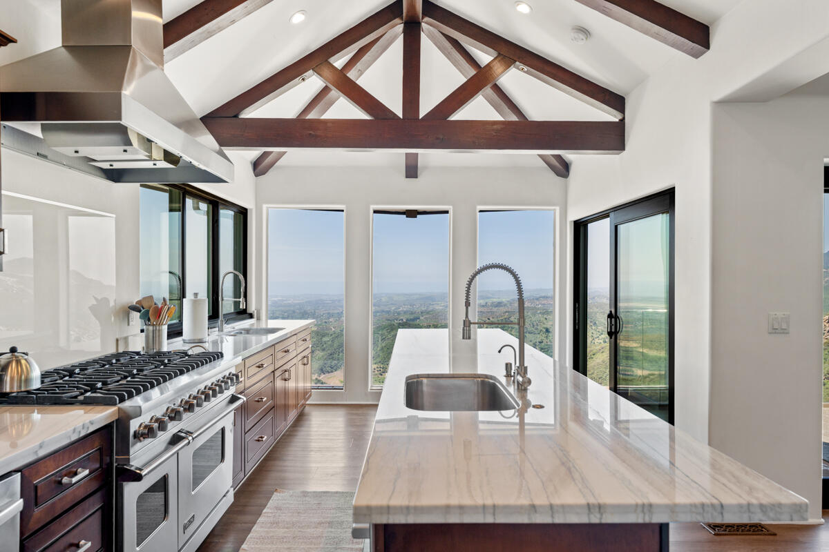 Modern kitchen with a marble island, exposed timber beams, and windows overlooking hills and ocean.
