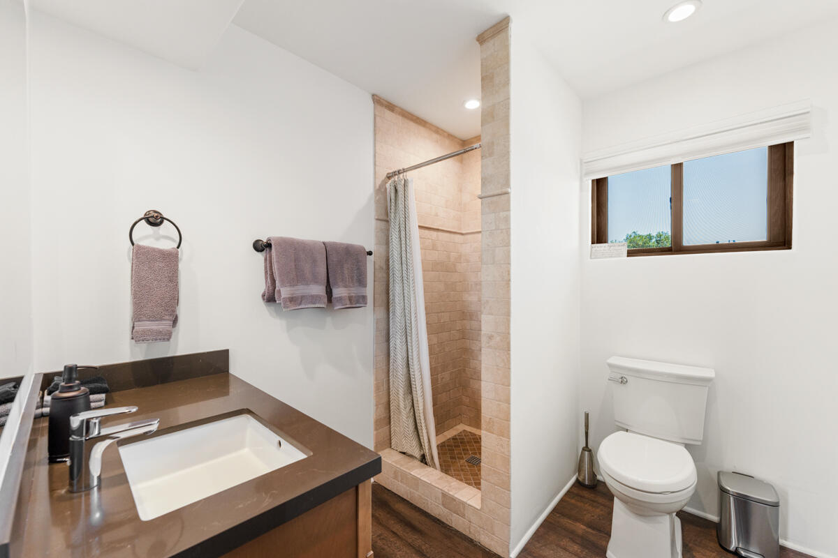 Bathroom with a beige tiled walk-in shower, dark countertop vanity, and toilet beneath a window.