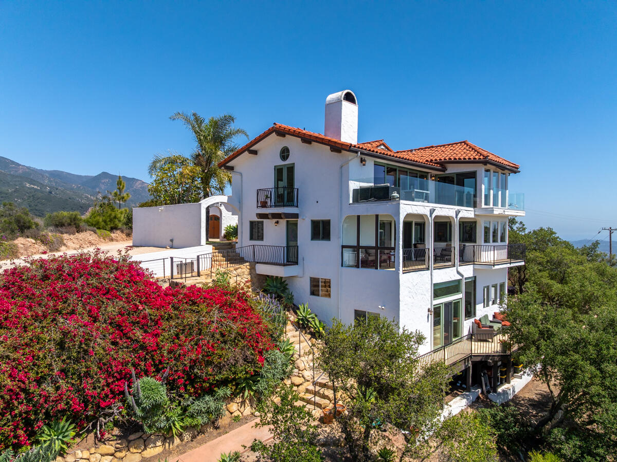 White multi-level home with terracotta roof, glass balconies, and flowering hillside landscaping.