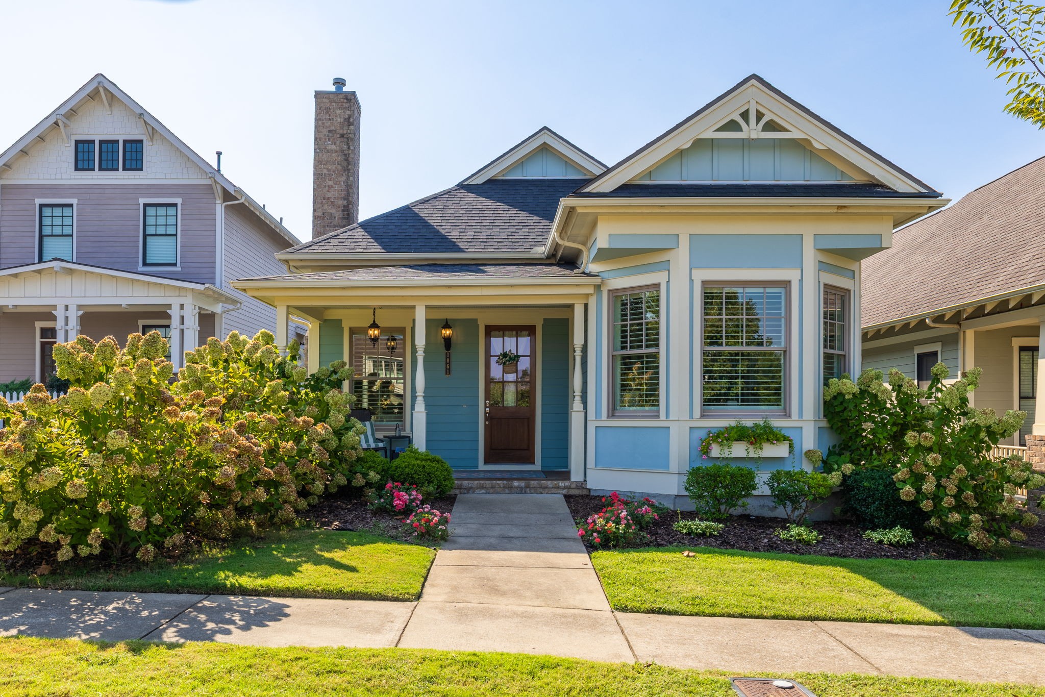 Blue house with a covered porch, bay window, and lush flower beds along the front walkway.