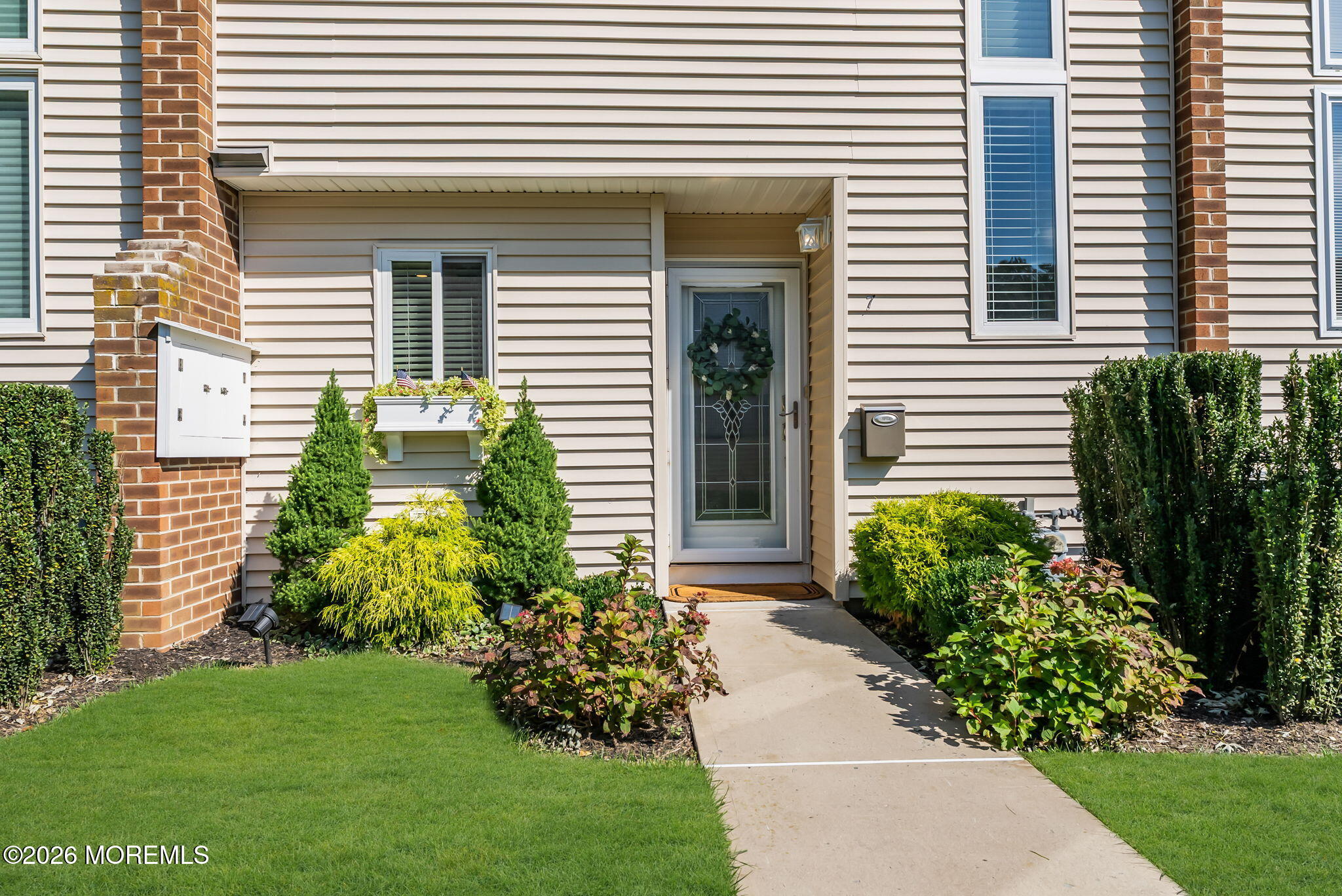 Home entrance with brick accents, a wreath on the glass door, landscaped shrubs, and a concrete walkway.