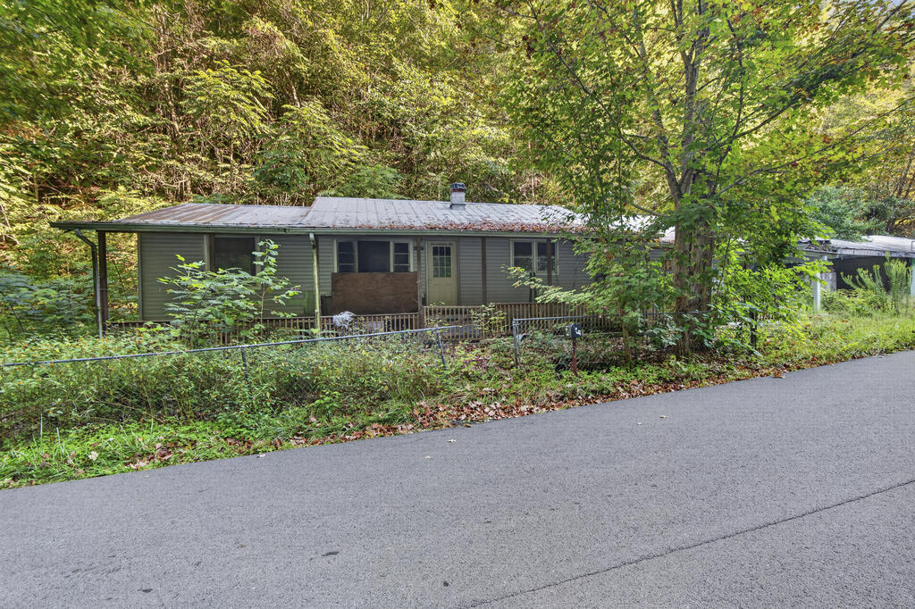 Single-story gray house with a metal roof behind a fence, surrounded by trees beside a paved road.