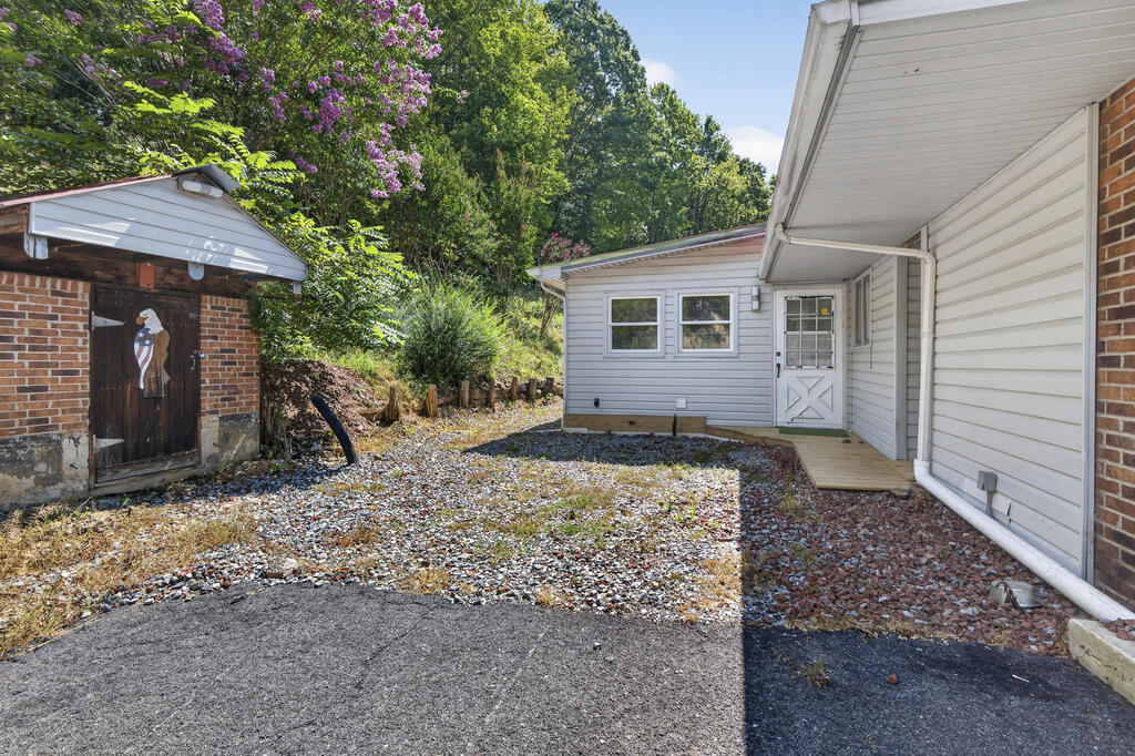 House and brick outbuilding around a gravel courtyard, with leafy trees and purple blossoms behind them.