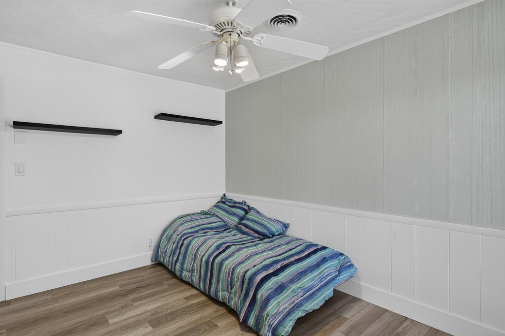 Bedroom with blue striped bedding, white wall paneling, a ceiling fan, and wood-look flooring.