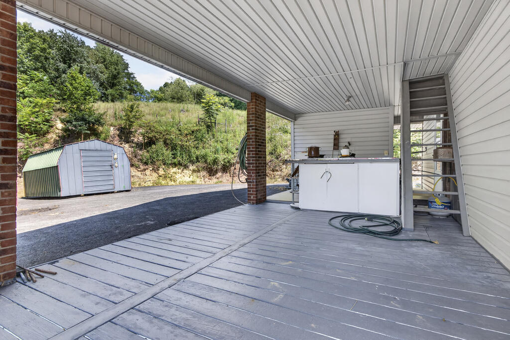 Covered porch with gray plank flooring, brick supports, and a utility counter beside a wooded hillside.