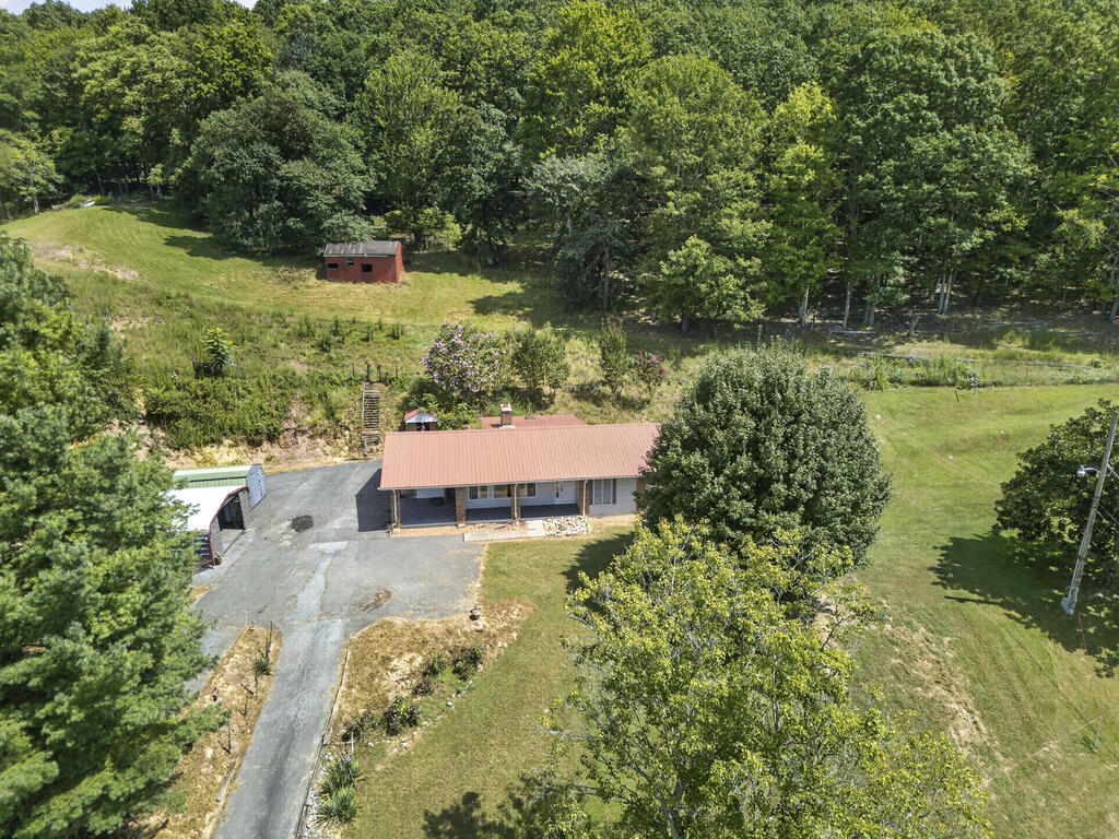 Aerial view of a single-story home with a red metal roof, driveway, and wooded grounds.