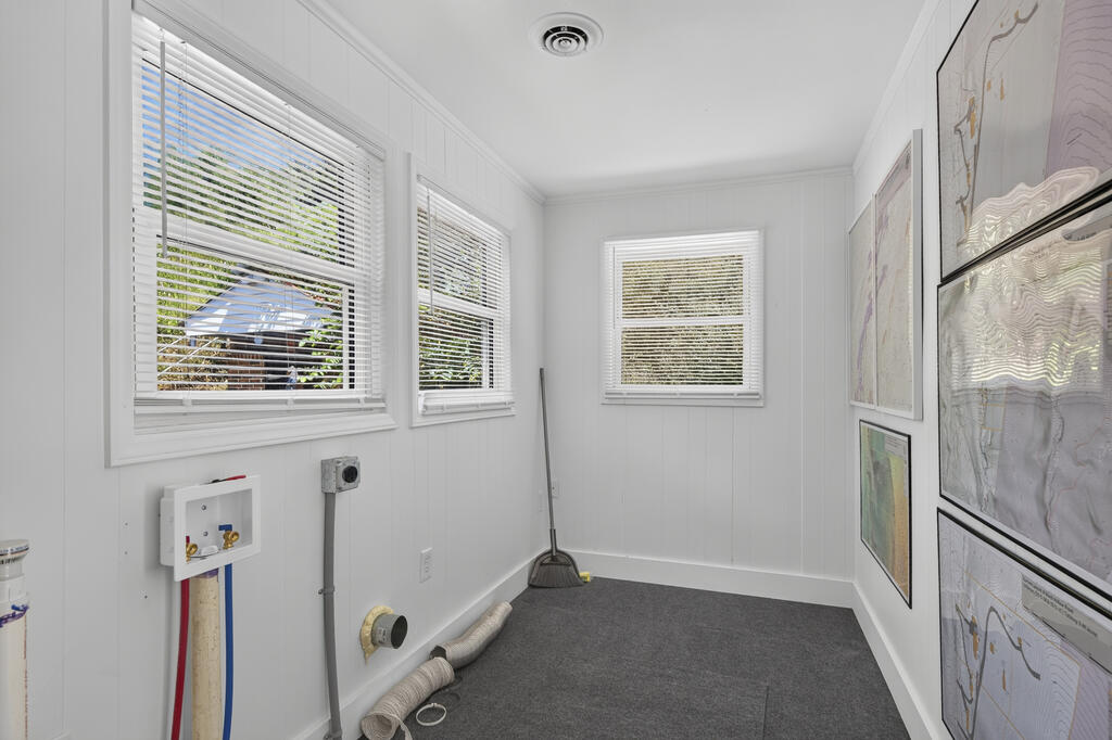 Vacant utility room with gray carpet, three windows, plumbing connections, and framed maps.