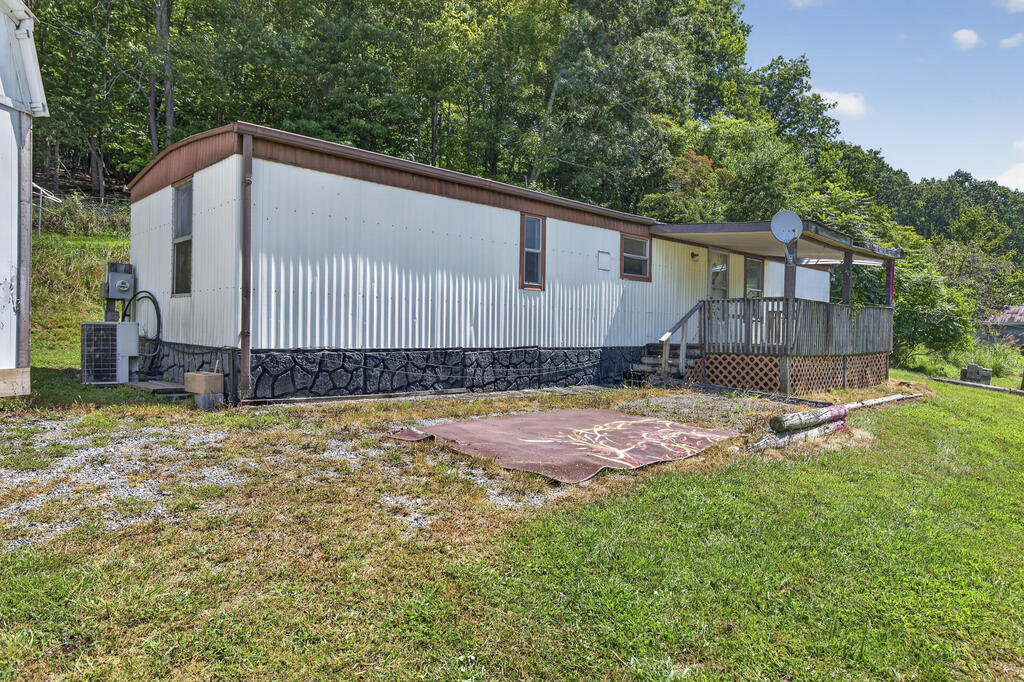 White manufactured home with a covered wooden porch, gravel yard, and mature trees behind it.