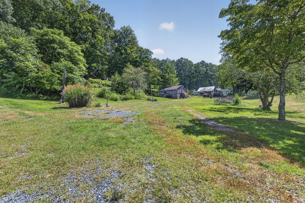 Rural property yard with mature trees, gravel patches, and several outbuildings in the distance.