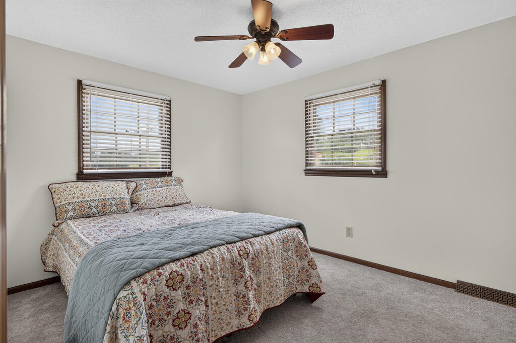 Bedroom with a patterned bed, two windows with blinds, and a ceiling fan.