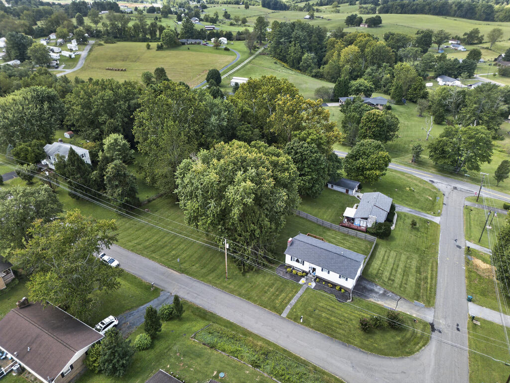 Aerial view of a white single-story house with a broad lawn beside a road intersection and wooded fields.