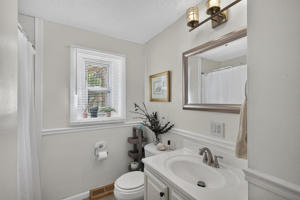 Bathroom with white vanity and oval sink, toilet, shower curtain, and window with blinds.