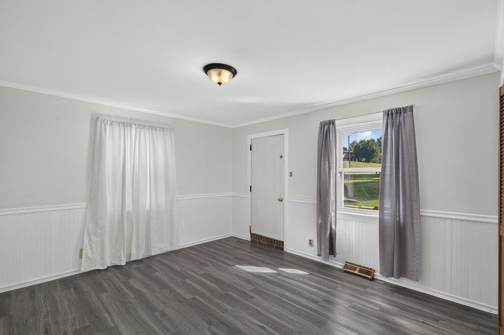 Empty room with gray plank flooring, white beadboard wainscoting, and a curtained window overlooking a lawn.
