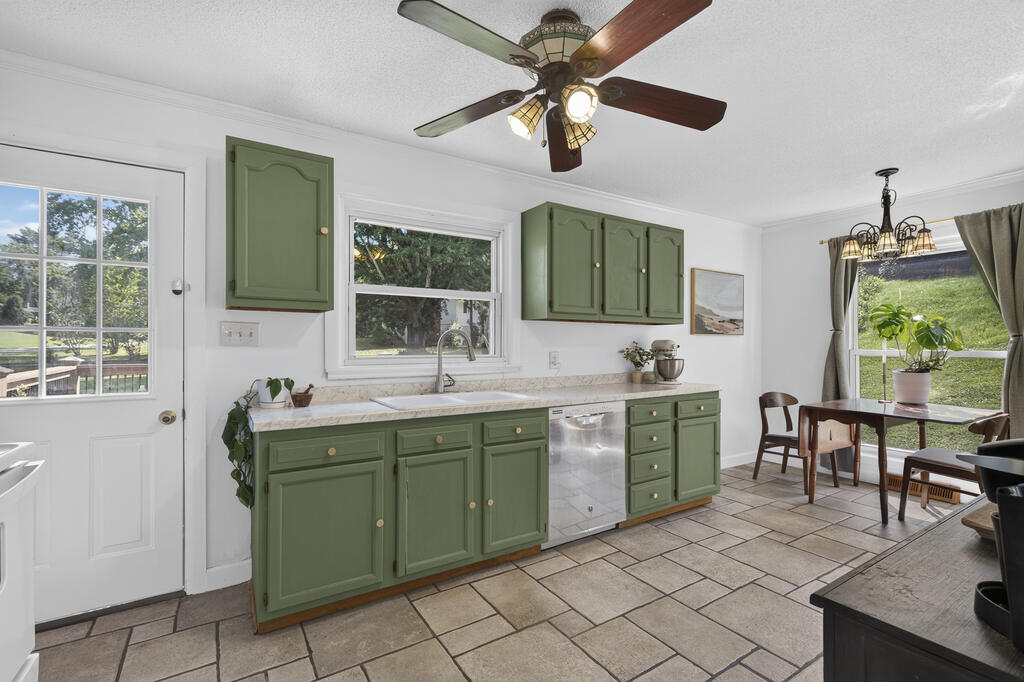 Kitchen with sage-green cabinets, tiled floors, stainless-steel dishwasher, and a dining area beside large windows.