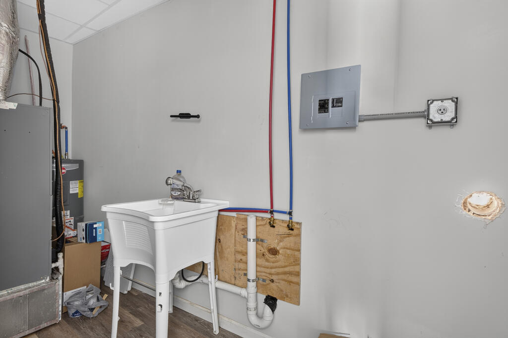 Utility room with a laundry sink, exposed plumbing, electrical panel, and water heater.