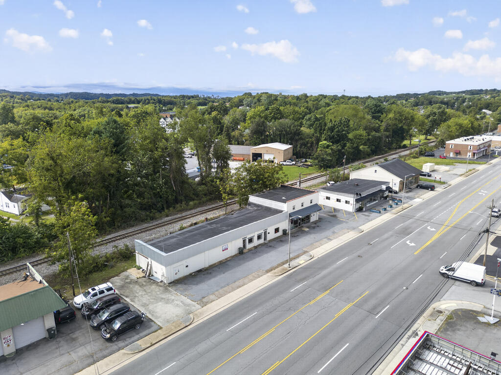 Aerial view of commercial buildings beside a main road, with railroad tracks and wooded surroundings.