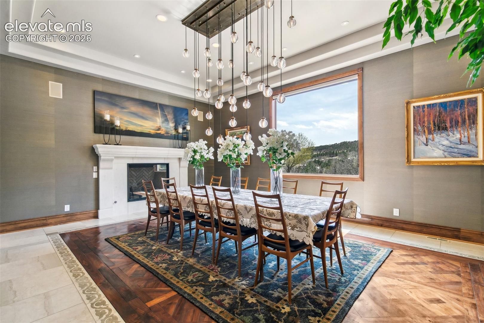 Dining room with a long table, white fireplace, pendant lights, and a large window overlooking wooded hills.