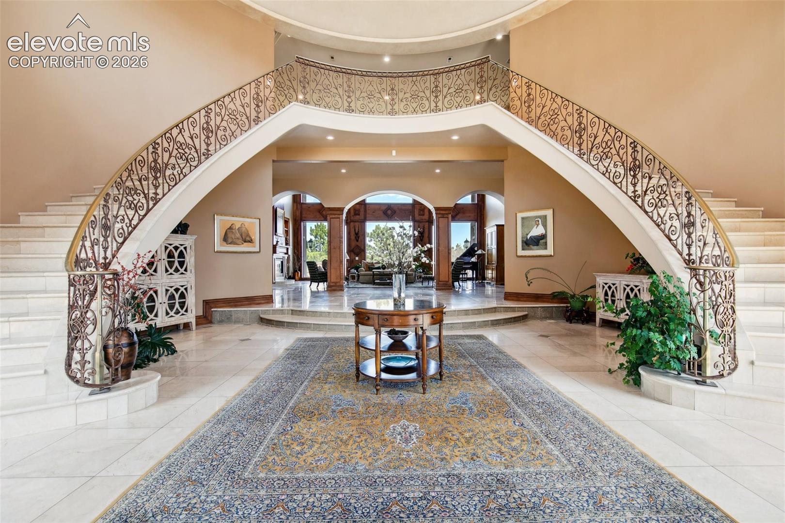 Grand foyer with a sweeping curved staircase, ornate iron railings, marble floors, and a patterned rug.