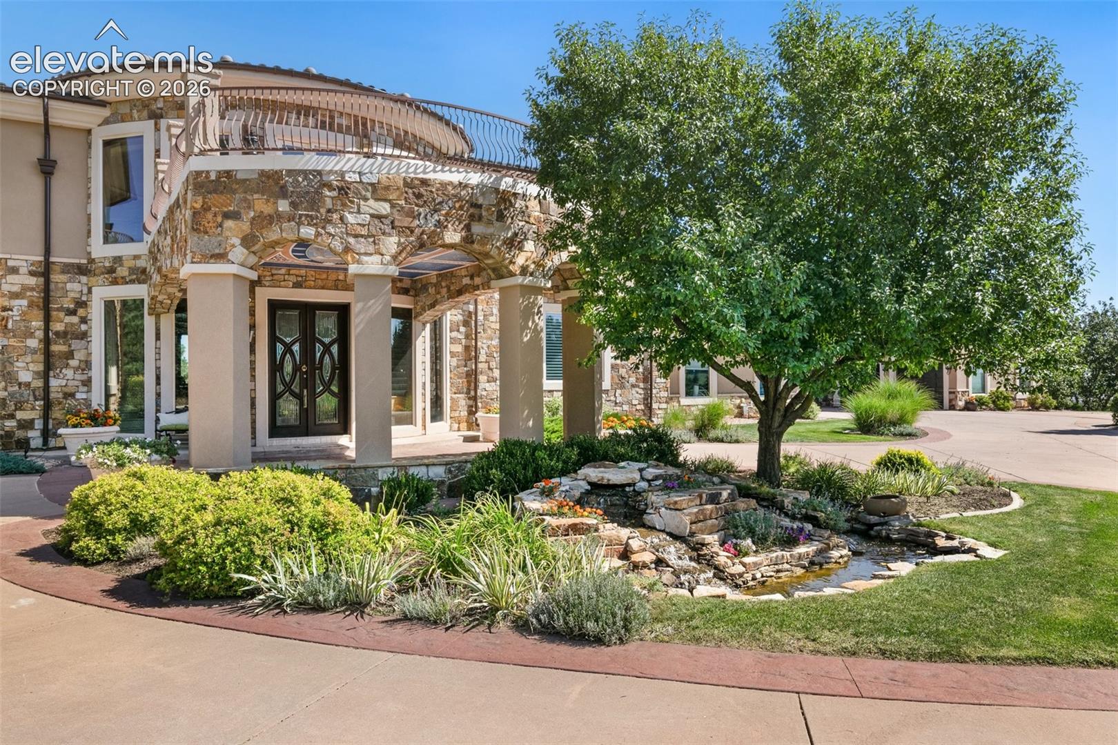 Stone-and-stucco home entrance with a curved balcony and landscaped garden with a small water feature.
