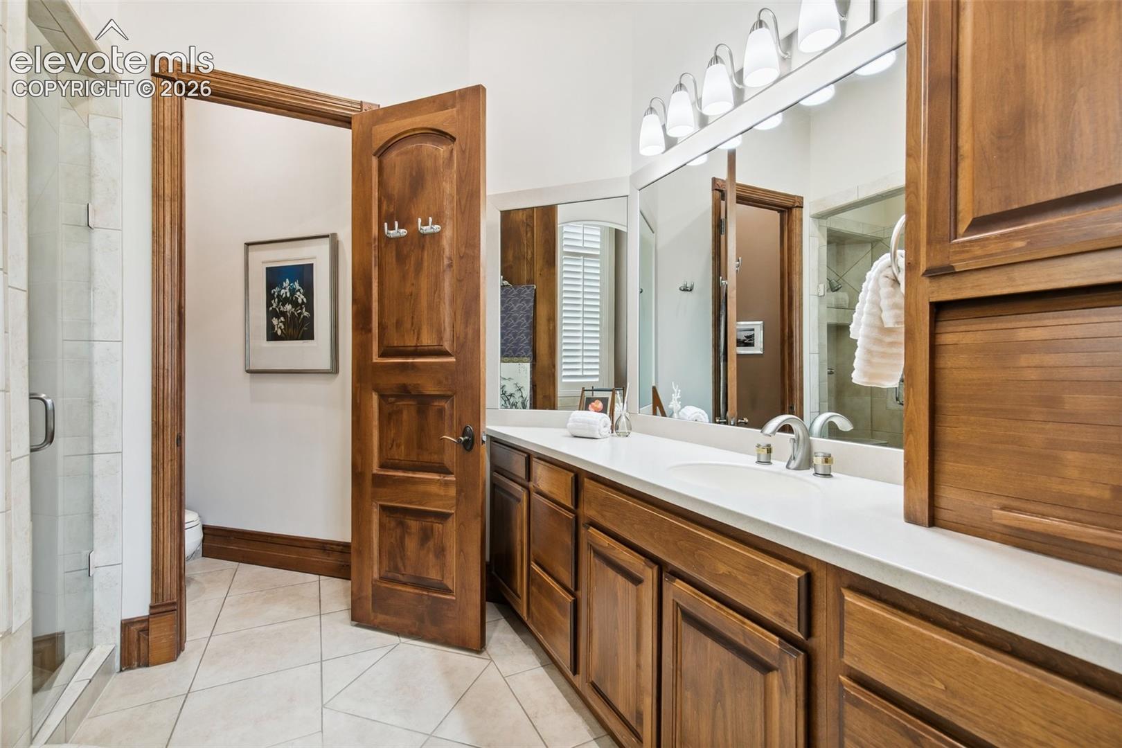 Bathroom with a long white vanity, warm wood cabinetry, large mirrors, and a tiled glass shower.