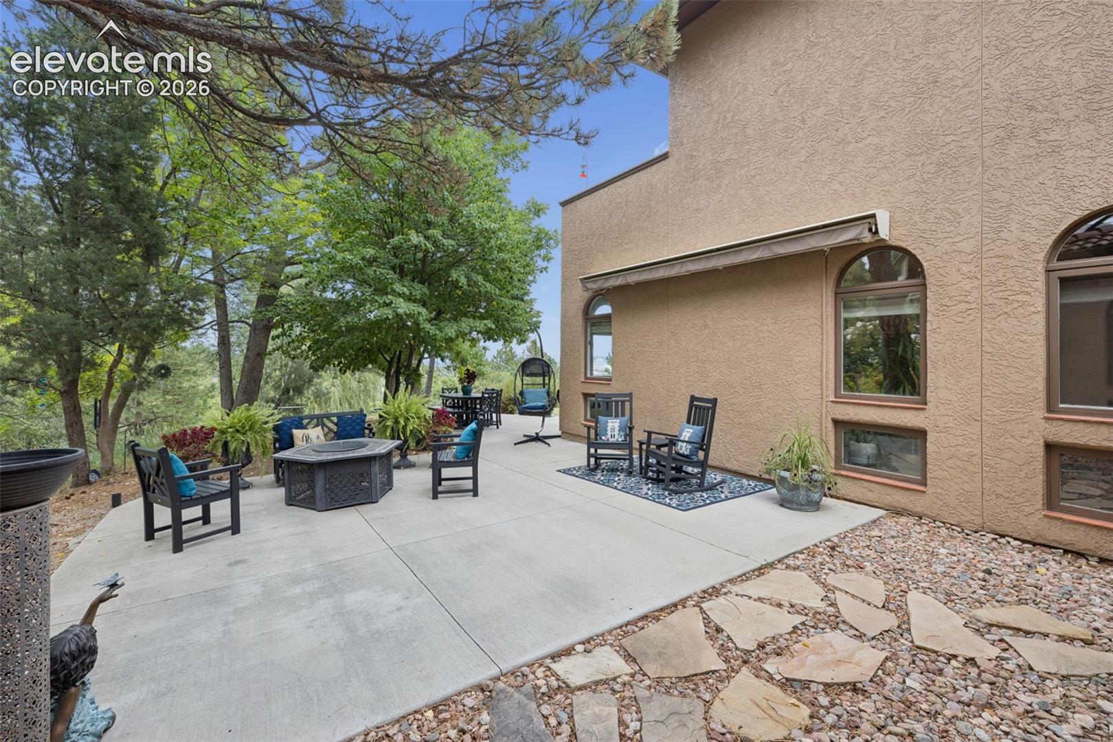 Furnished concrete patio beside a tan stucco home, shaded by mature trees.